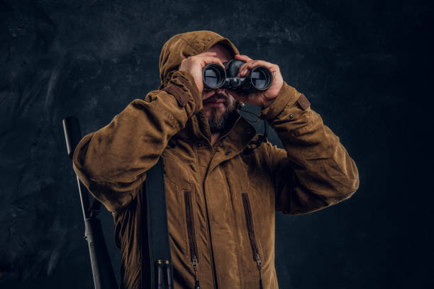 istock Hunter with shotgun watching through binoculars. Studio photo against dark wall background 1182682356