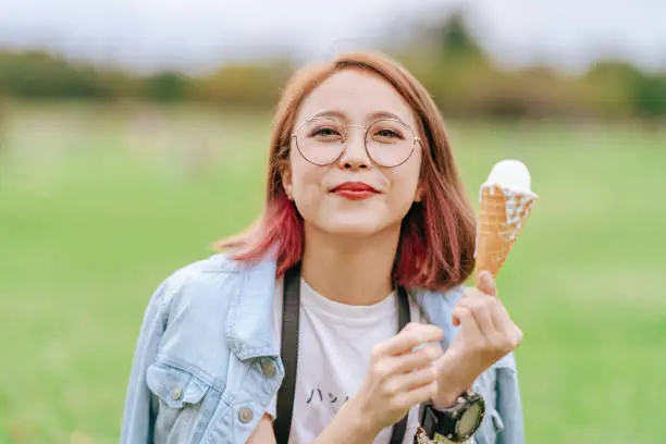 A happy young woman is sitting on grass and eating ice cream happily in a public park.