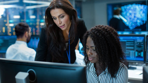 istock Female Team Leader Consults Young Computer Engineer. They Work in a Crowded Office on a Neural Network/ Artificial Intelligence Project. 1182697689