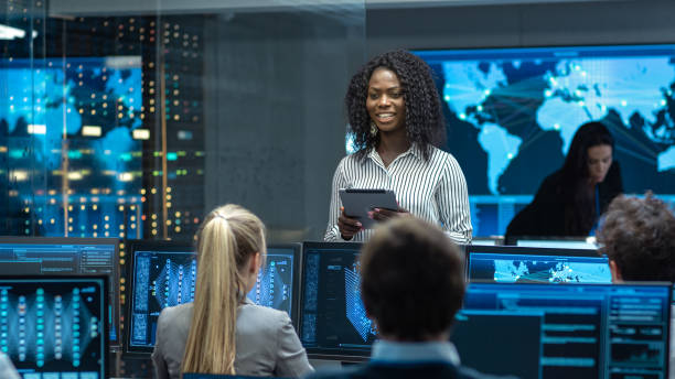 istock Chief Project Engineer Consults Tablet Computer and Holds Briefing for a Team of Scientists that are Building Machine Learning System. Displays Show Working Model of Neural Network. 1182697709