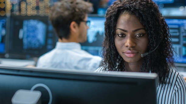 il rappresentante del servizio clienti femminile risponde alle domande del cliente in un auricolare. lavora per una grande azienda tecnologica. office ha un team multietnico di specialisti, display mostrano infografiche utili - mittente foto e immagini stock