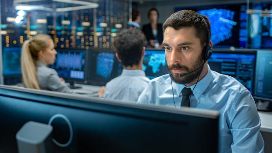 Customer Service Representative Answer Client's Questions in a Headset. He Works for a Big Technological Company. Office Has Multi-Ethnic Team of Specialists, Displays Show Useful Information.