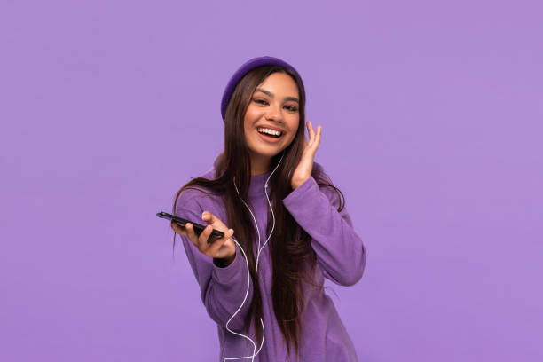 Pretty african-american girl in a hat and sweater dancing listening to music on a mobile phone with wired headphones isolated over purple background. Pretty african-american girl in a hat and sweater dancing listening to music on a mobile phone with wired headphones isolated over purple background. Enjoying life. headphones plugged in photos stock pictures, royalty-free photos & images