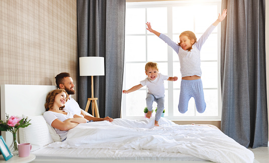 happy family mother, father and children laughing, playing and jumping in bed in bedroom at home