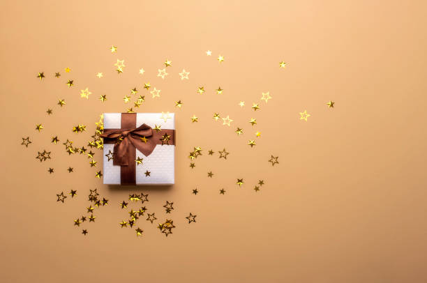 Gift box and shiny star-shaped confetti. Christmas gifting frame White Christmas gift box with a brown bow ribbon and golden confetti, star-shaped on a tan-colored background. Christmas gifting concept. Festive gift gift lounge stock pictures, royalty-free photos & images