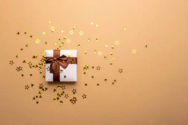White Christmas gift box with a brown bow ribbon and golden confetti, star-shaped on a tan-colored background. Christmas gifting concept. Festive gift