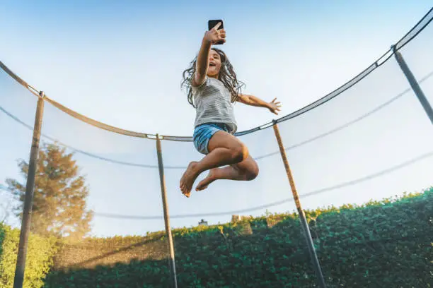 little girl jumping high on trampoline under blue sky while chatting on mobile