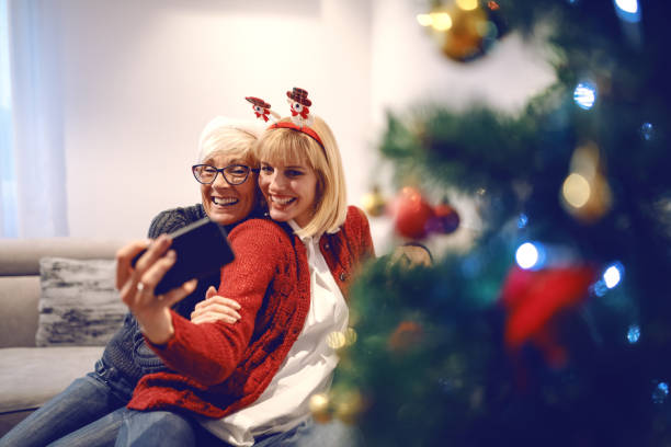 carina donna caucasica e sua madre che si fanno selfie mentre sono sedute sul divano in salotto. in primo piano c'è l'albero di natale. - senior adult winter senior women daughter foto e immagini stock