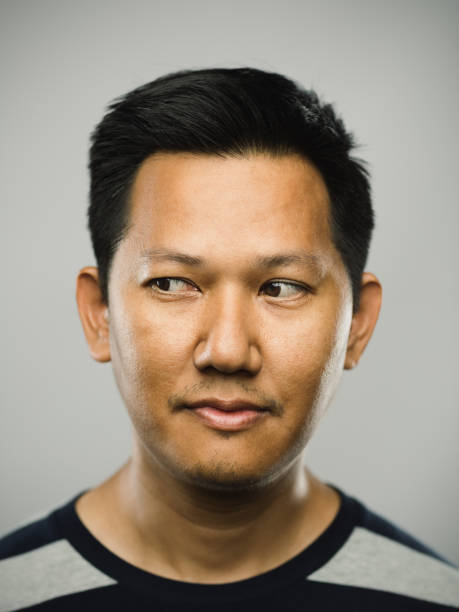 Real malaysian man with blank expression looking to the side Close up portrait of asian man with blank expression looking to the side against gray white background. Vertical shot of malaysian real people observing in studio with black hair and modern haircut. Photography from a DSLR camera. Sharp focus on eyes. sideways glance stock pictures, royalty-free photos & images