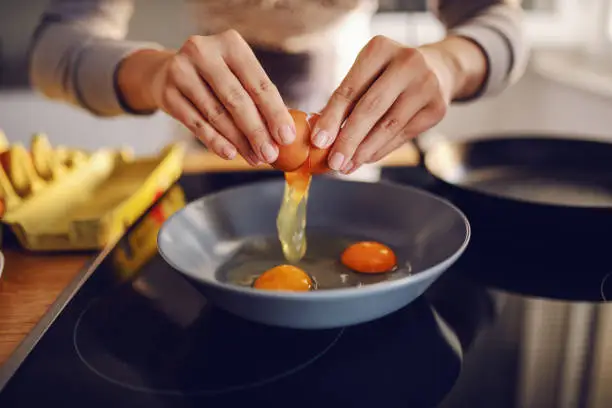 Close up of caucasian woman breaking egg and making sunny side up eggs. Domestic kitchen interior. Breakfast preparation.