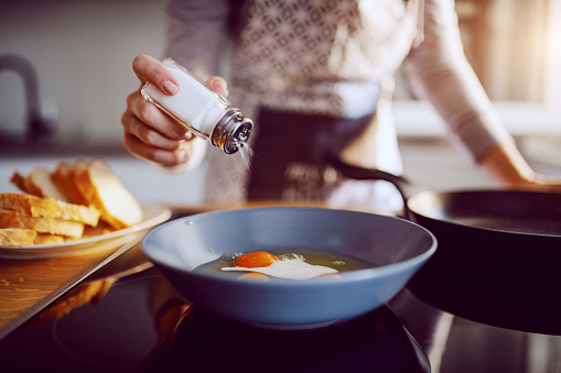 Primer plano de la mujer caucásica añadiendo sal en los huevos soleados mientras está de pie en la cocina junto a la estufa. photo
