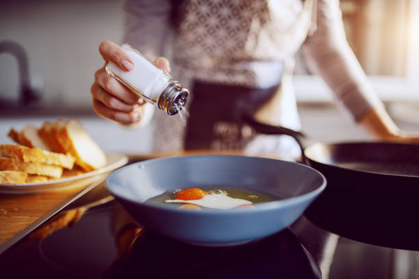primer plano de la mujer caucásica añadiendo sal en los huevos soleados mientras está de pie en la cocina junto a la estufa. - sales fotografías e imágenes de stock