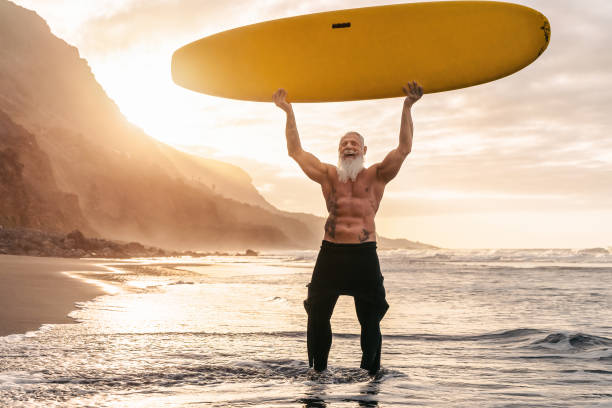 счастливый подходят старший весело серфинга на закате время - спортивный бородатый человек подготовки с доской для серфинга на пляже - пожи - surfing surf wave extreme sports стоковые фото и изображения