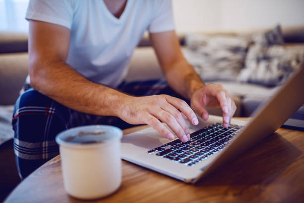 Cropped photo of caucasian man in pajamas typing on laptop keyboard and surfing the internet while sitting on sofa in living room in morning. Selective focus on hands. Cropped photo of caucasian man in pajamas typing on laptop keyboard and surfing the internet while sitting on sofa in living room in morning. Selective focus on hands. arabica coffee drink photos stock pictures, royalty-free photos & images