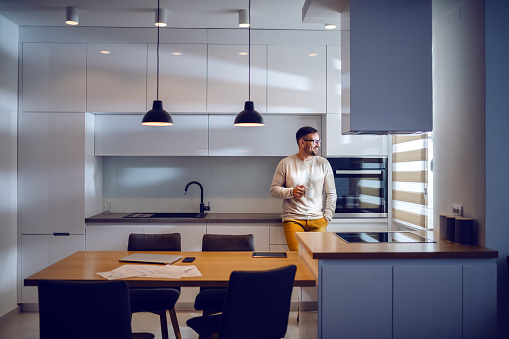 Full length of handsome Caucasian man dressed casual leaning on kitchen counter, drinking coffee and looking trough window. Modern kitchen interior.