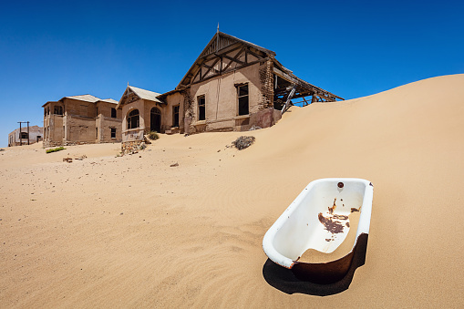 Bizarre surreal scene in the Namibian Desert. Old broken bathtub on Sand Dune in front of abandoned half-timbered old historic buildings in Ghost Town Old Diamond Mine under deep blue desert sky. Kolmanskop, Luderitz, Namibia, Africa.