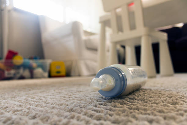 Low angle view of a feeding bottle with some milk left on white carpet with some toys and a toddler's chair in the background.