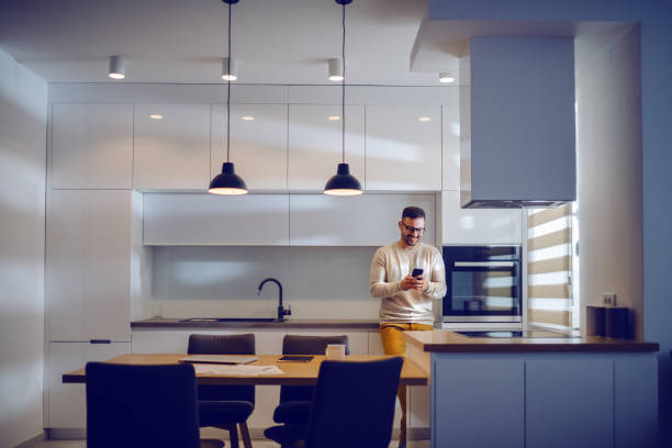 istock Attractive Caucasian smiling man dressed casual leaning on kitchen counter and using smart phone to send a message. Home interior. 1182729008