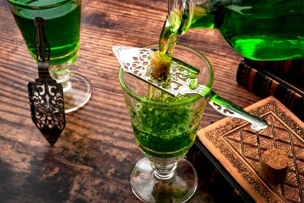 Photo of Alcoholic drink, creative stimulant and bohemian lifestyle concept theme with a vintage glass bottle pouring absinthe over a sugar cube in a stainless steel spoon next to books on a wooden table