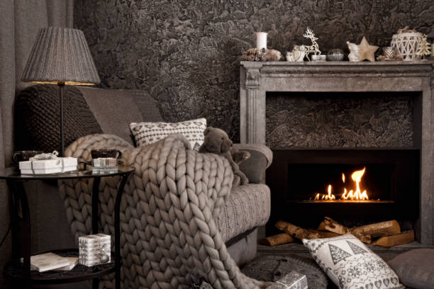 Modern cozy interior in gray shades with an armchair in front of fireplace. Modern cozy interior in gray shades with an armchair in front of fireplace. On the chair there are nice knitted blanket, cushions and Teddy bear. A lamp and gifts on the table. Candle and other Christmas decorations standing above the fireplace. fireplace and armchair stock pictures, royalty-free photos & images