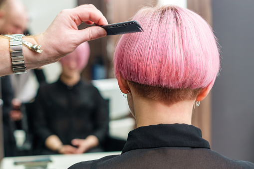 The hairdresser is using comb for female pink hair at beauty salon. Male hand is combing woman short pink hair in hair salon.