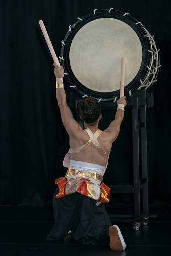 Taiko drummer hits the big drum on stage on a black background, back view.