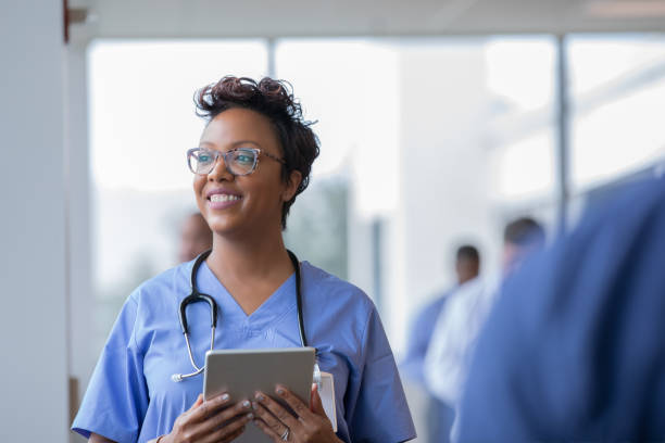 female nurse or doctor smiles while staring out window in hospital hallway and holding digital tablet with electronic patient file - doctor stethoscope healthcare worker professional occupation imagens e fotografias de stock