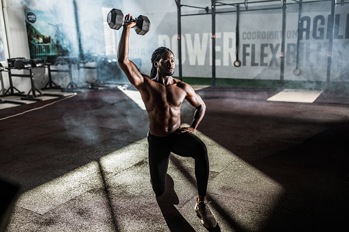 Young African American athlete exercising strength with dumbbell while being in lunge position at gym.