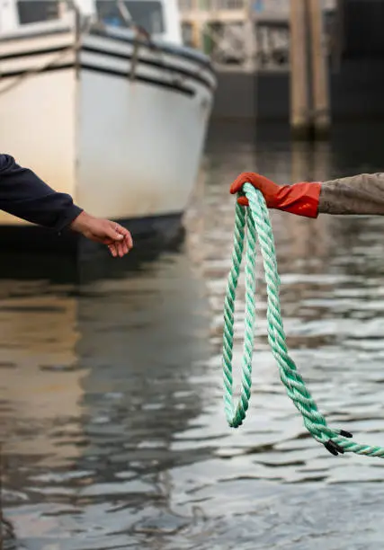 A photo with two hands in it - one is wearing an orange glove and holding a thick coiled green and white rope that is used to tie up a boat. They are handing the rope to another bare hand that is reaching out over water, with a white boat docked in the background.