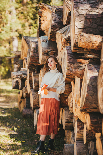 Beautiful woman with autumn maple leaves posing in the background of wood logs in forest. Vertical view