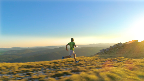 AERIAL, LENS FLARE: Flying behind athletic young man sprinting down a grassy hill at sunset. Cinematic shot of fit male fell running in the scenic mountains in Slovenia on a sunny summer evening.