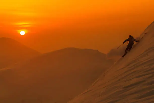 SILHOUETTE: Spectacular shot of pro skier riding off trail on a picturesque sunny evening in Niseko. Unrecognizable male tourist shredding the powder snow covering the beautiful mountains in Japan.