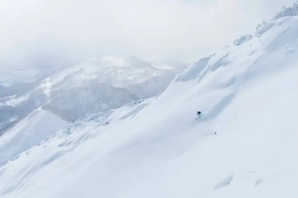 AERIAL: Unrecognizable man shredding the snowy mountain terrain while on fun ski trip in Japan. Flying above extreme skier riding off trail and through the fresh powder snow in picturesque Niseko.