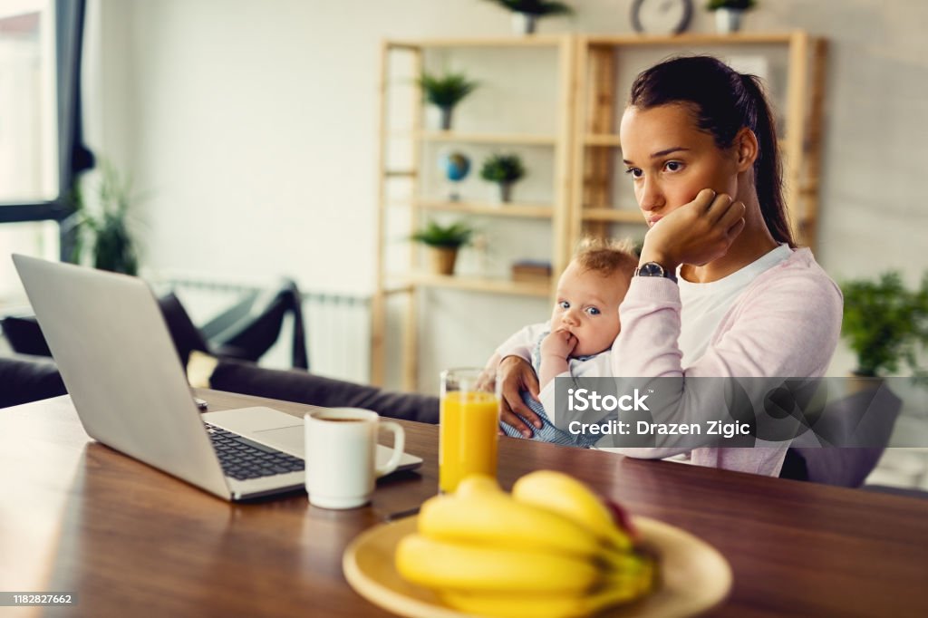 Young distraught mother thinking of something while being with her baby at home. Young woman with baby experiencing postpartum depression and contemplating at home. Mother Stock Photo