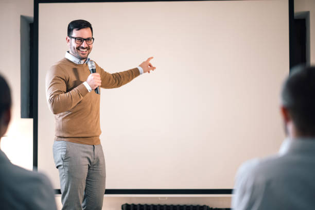 istock Portrait of a speaker on a seminar, talking on microphone and pointing at blank screen. 1182838369