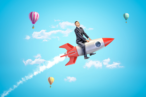 Young businessman riding toy rocket in blue sky with hot air balloons in background. Nothing is impossible. Successful career move. Investment in space exploration.