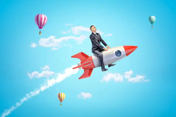 Photo of Young businessman riding toy rocket in blue sky with hot air balloons in background.