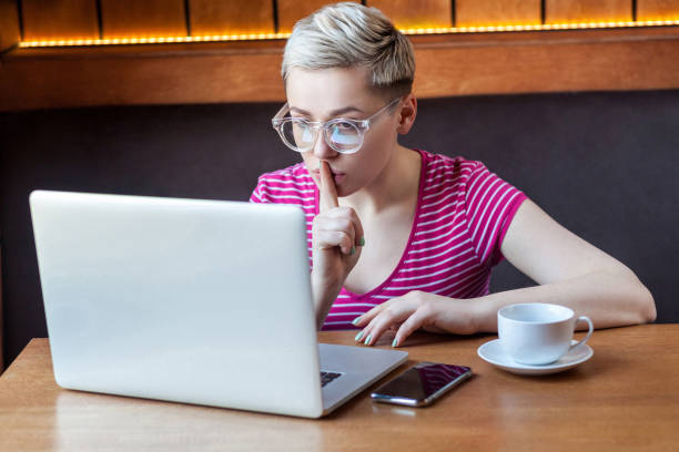 seien sie ruhig bitte! junge mädchen freiberufler mit blonden kurzen haaren, in rosa hemd und brille sitzt im café und macht videoanruf auf laptop, schlanker finger auf mund zeigt stille, - silence finger on lips businesswoman one person stock-fotos und bilder