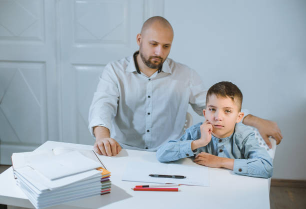 istock Sad kid sitting at a table next to his father, who is angry at him. 1182843301