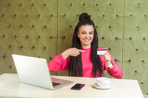 Portrait of beautiful happy young businesswoman with black dreadlocks hairstyle in pink blouse is sitting in cafe, pointing finger to credit card, easy to order online buy, looking at camera. Indoor