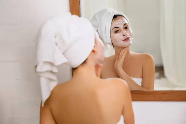 Woman with towel is applying mask on her face in front of the mirror at home. Skin care.