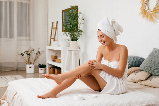 woman applying cream on her legs Young beautiful woman in towel applying cream on her legs after shower at home moisturizer stock pictures, royalty-free photos & images