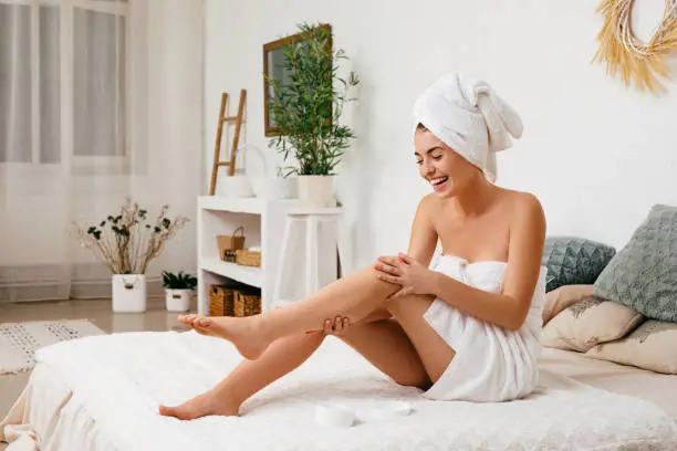Young beautiful woman in towel applying cream on her legs after shower at home