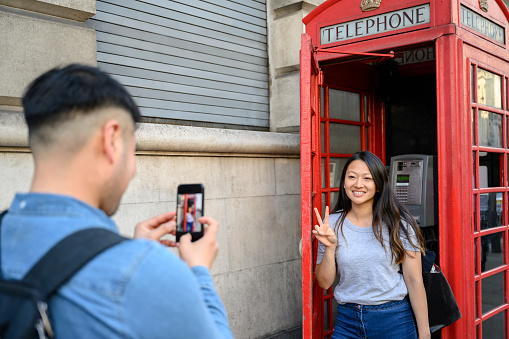 Male Chinese vacationer taking smart phone photo of young female friend posing in iconic London red telephone box and making peace sign gesture.