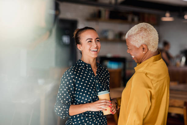 Enjoying Their Breaks Together Two women colleagues laughing while standing in a cafe at their workplace. One of the women is holding a take out hot drink cup. talking stock pictures, royalty-free photos & images