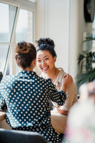 Two Friends Talking at Work Two women colleagues laughing while standing near a window together at their workplace. There is a laptop on the table in front of them. face to face stock pictures, royalty-free photos & images