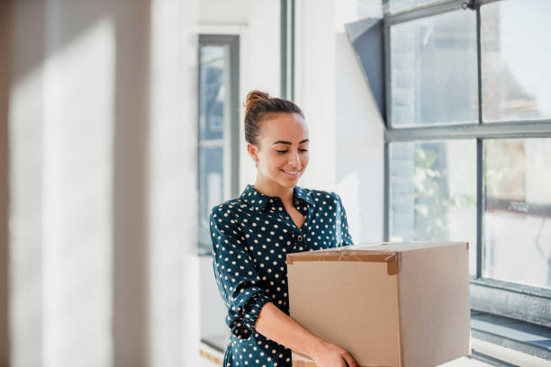 Unloading New Office Supplies Woman laughing and looking down while carrying a cardboard box at work. carrying stock pictures, royalty-free photos & images