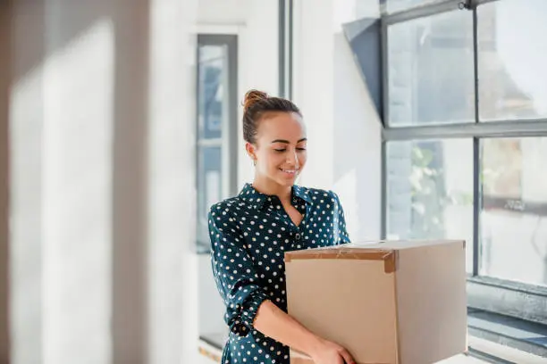 Woman laughing and looking down while carrying a cardboard box at work.