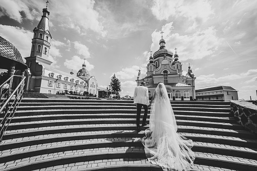 Bride and groom go to church on wedding day. Wedding couple holding hands and going up the stairs of the castle. Back view. Black and white photo.