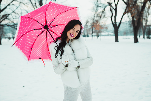 Beautiful smiling young woman holding pink umbrella on a snowy winter day in the public park outdoors